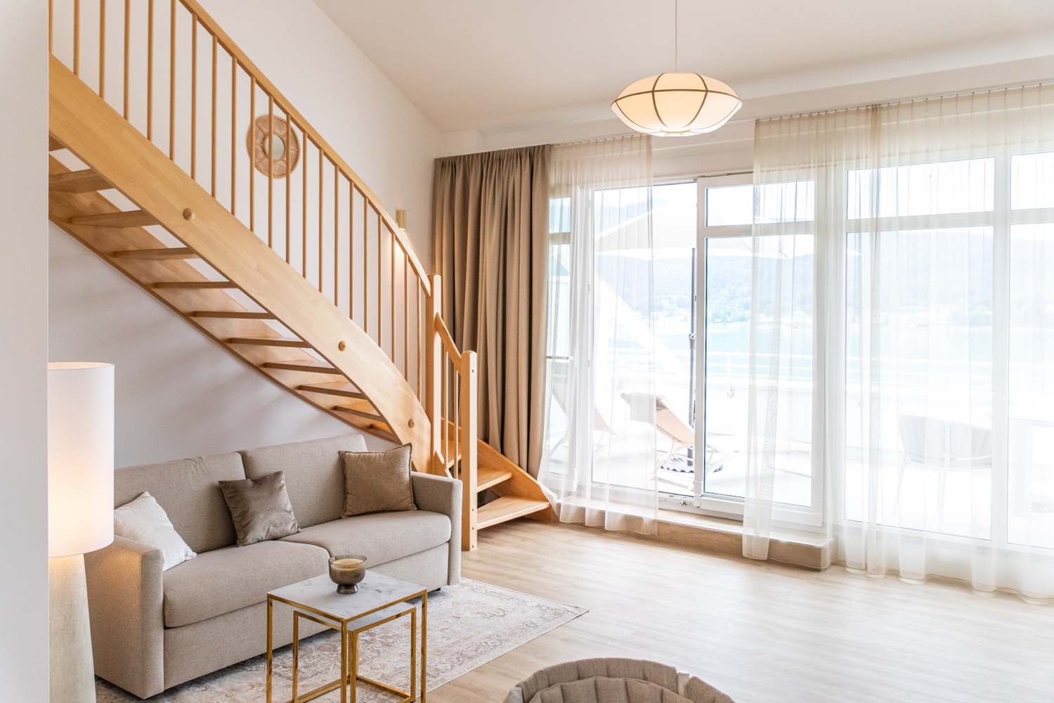 Heller Wohnraum mit Holz-Galerietreppe, Sofa und großen Fenstern mit Seeblick